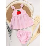 A baby girl's pink and white striped summer outfit featuring a red apple applique on the chest and ruffle sleeve top, paired with matching shorts, laid flat on a woven mat.