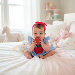 A cute baby girl wearing a blue and white striped summer outfit with a ruffle sleeve top and matching shorts, sitting on a bed with a red bow headband.