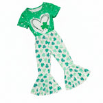 Green short sleeve girls t-shirt with a white heart and green clover print, paired with white flare pants covered in shamrock patterns, displayed alongside two small teddy bears and a white plush rabbit toy on a white background.